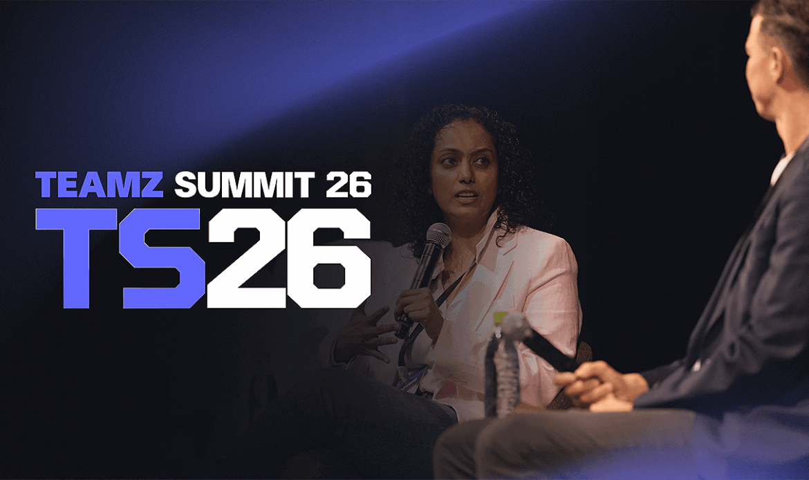 A woman speaking into a microphone onstage under a large lit background display reading SUMMIT 26.