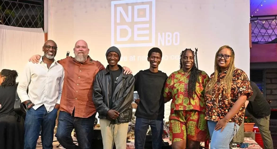 Group portrait photo of four smiling young innovators standing together shoulder-to-shoulder inside an office building.