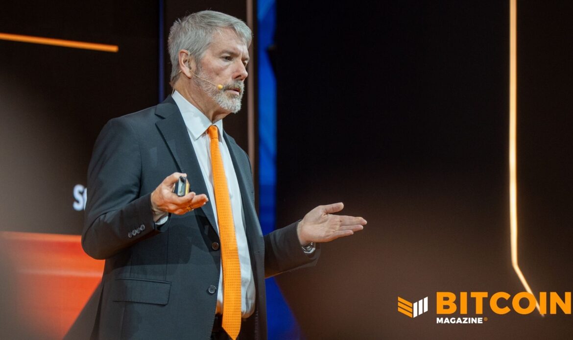 Michael Saylor speaking onstage in a dark suit with an orange tie against a dark background.