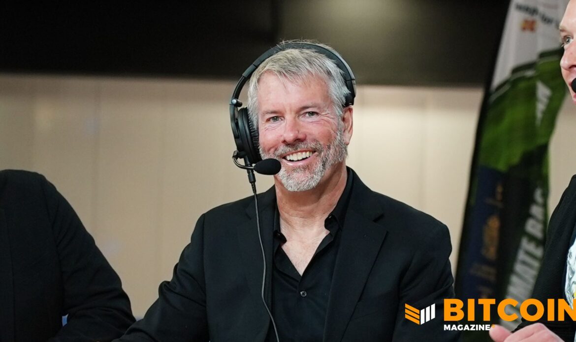 Headshot of a smiling Michael Saylor wearing a dark suit jacket and a broadcast headset microphone.