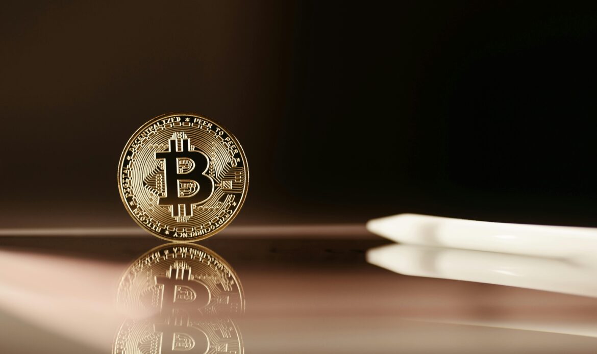 A small physical golden Bitcoin coin standing upright next to a blurry computer pointer pen